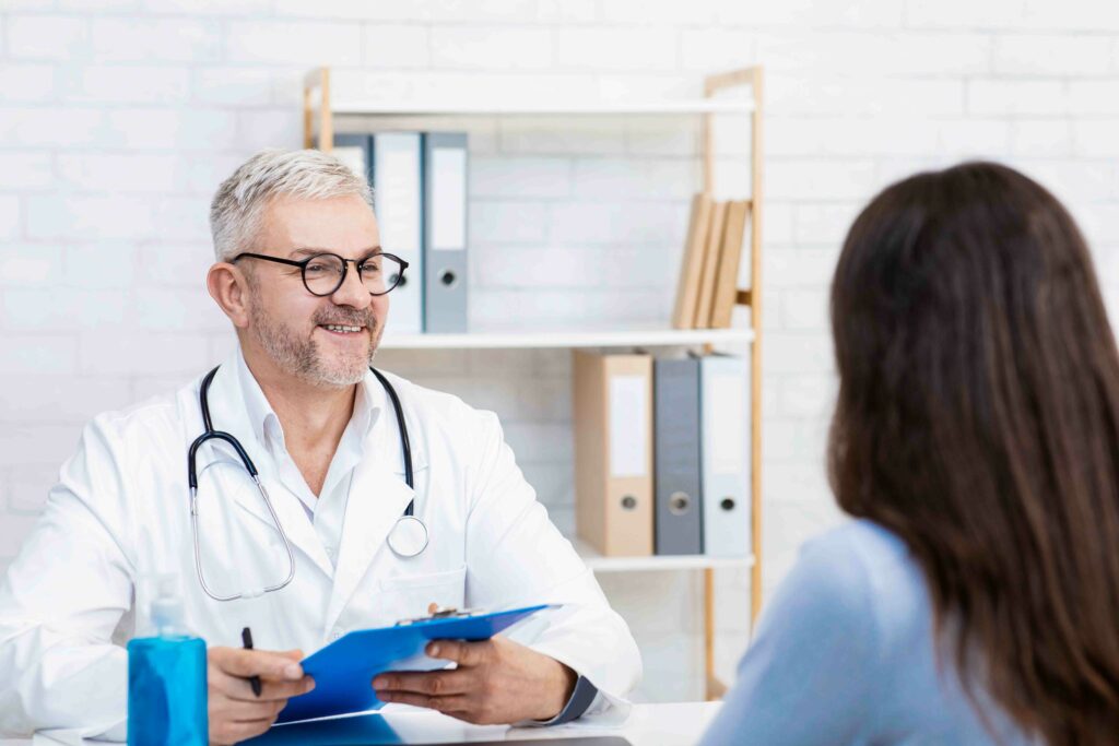 Family doctor visit, health care and decision problems in modern clinic. Smiling adult man in glasses and white coat talking with woman patient and notes symptoms in office interior, empty space