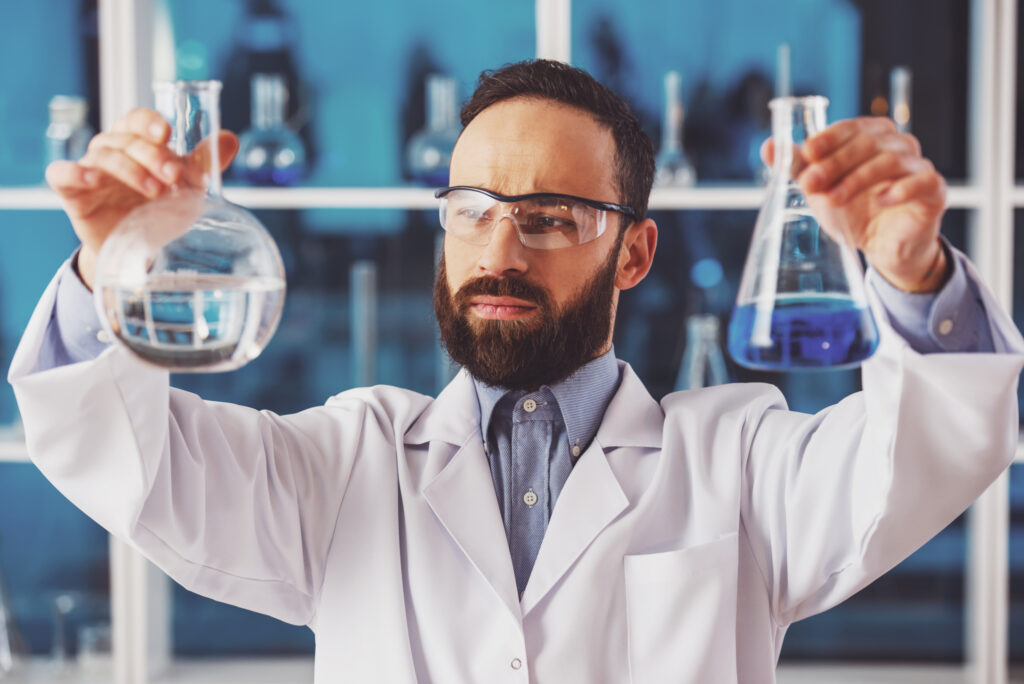 Handsome male scientist researcher in safety glasses observing liquid in flasks in a laboratory.