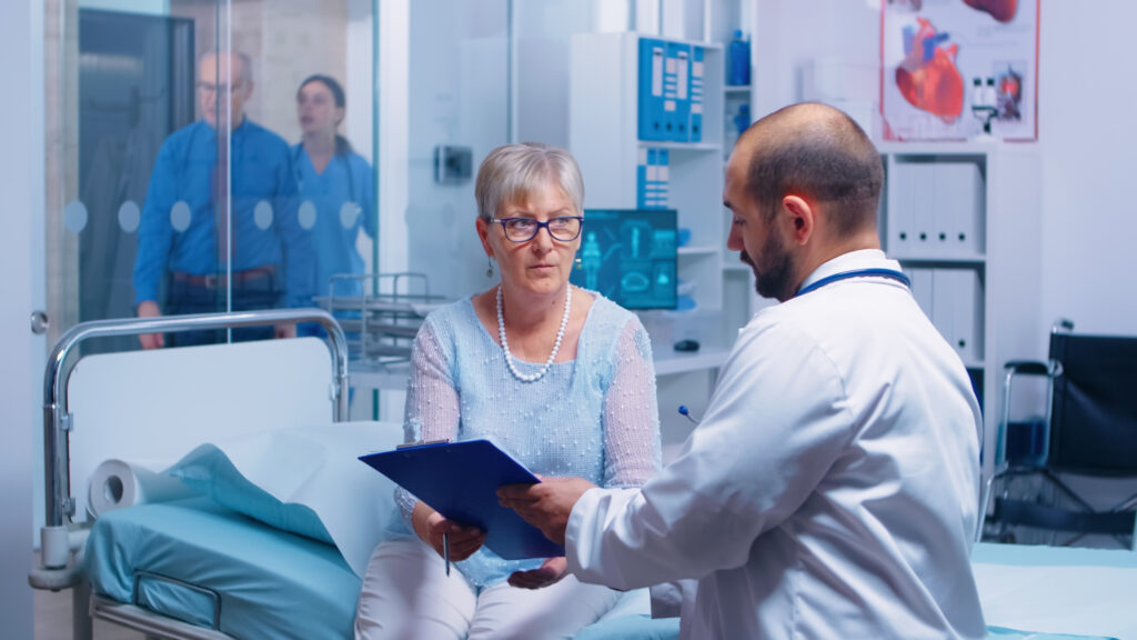 Senior patient signing medical decisions form sitting on hospital bed in modern private clinic. Doctor with clipboard, nurse working in backgorund. Healthcare medical medicinal system documents contract