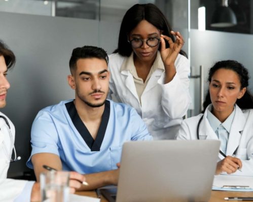 Professional multiracial team of doctors concentrated men and women in workwerar sitting at table, looking at notebook screen, taking notes, looking for solutions, clinic interior